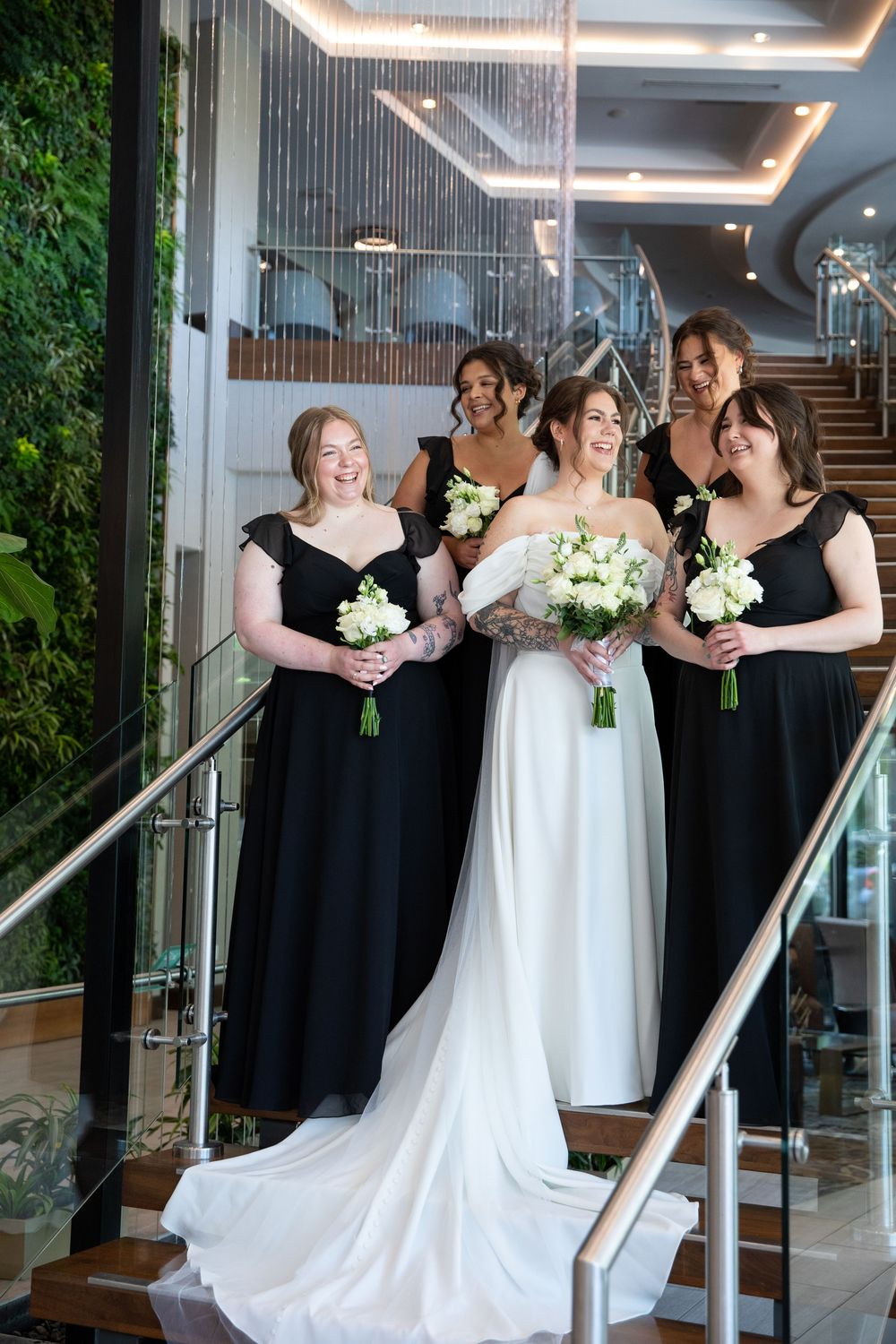 Bride in white gown with bridesmaids in black dresses holding white bouquets on elegant staircase.