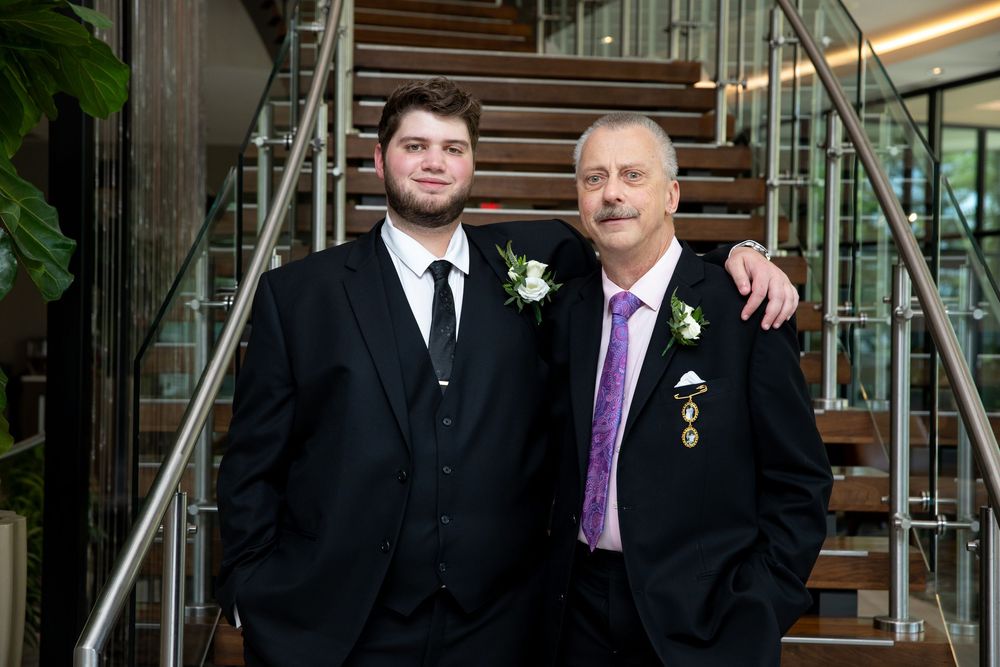Two men in formal black suits with boutonnieres pose together on a staircase at a wedding.