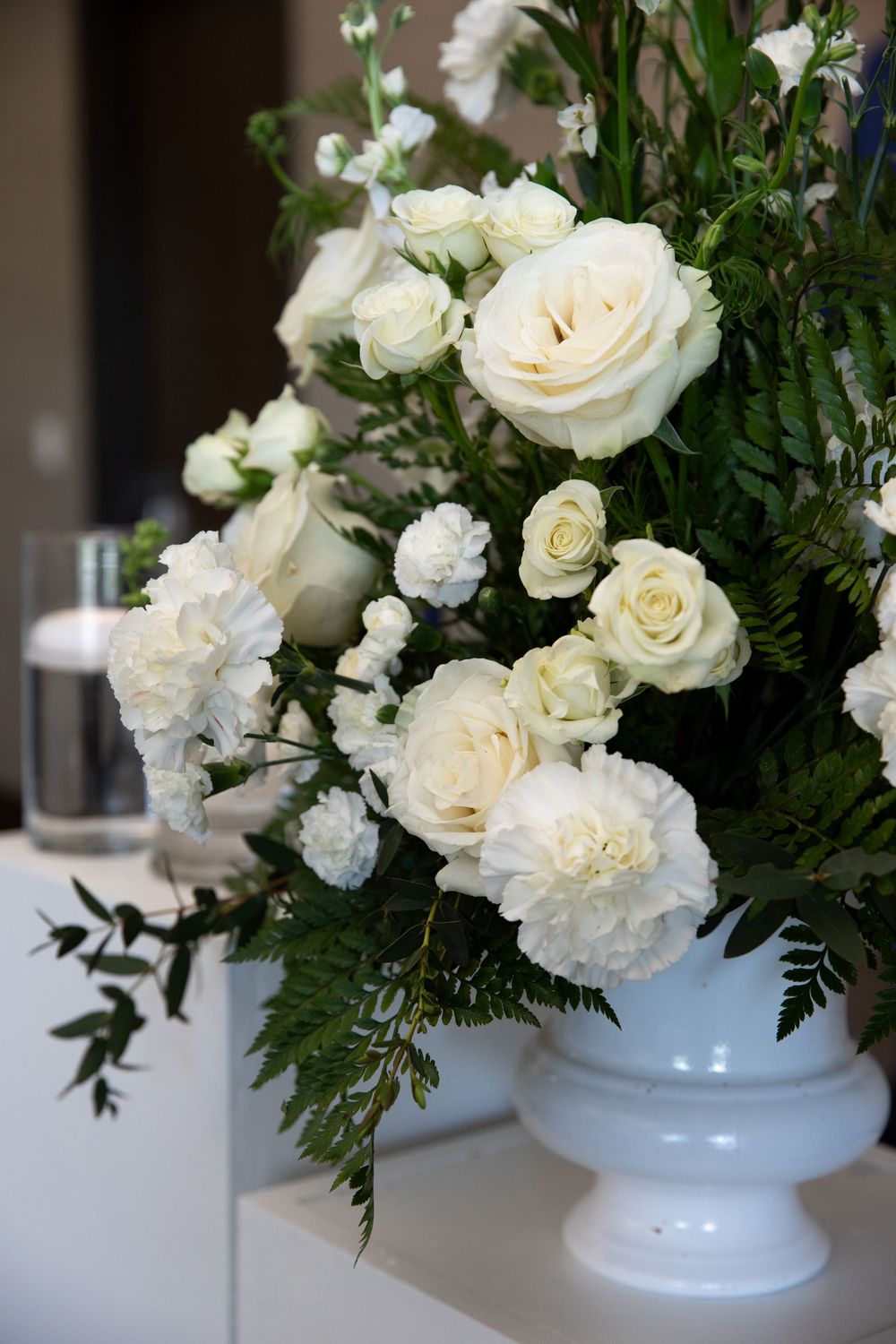 Elegant white rose and carnation wedding floral arrangement in a white ceramic urn vase with lush green ferns.