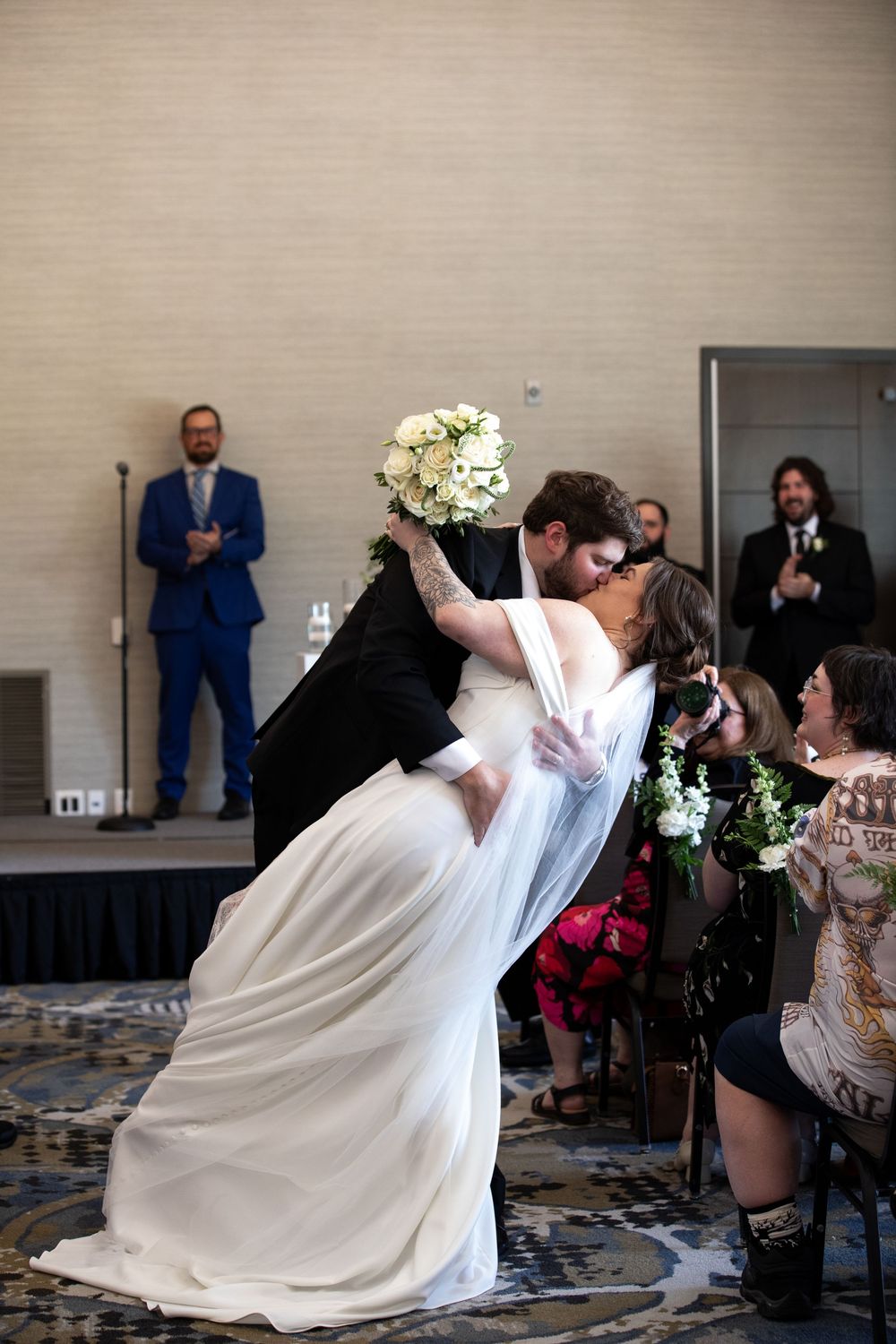 Bride and groom share a romantic dip kiss at their wedding ceremony while guests cheer around them.