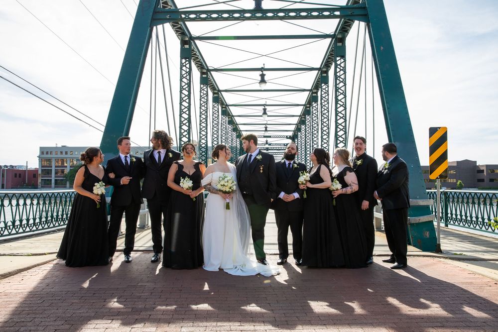 Wedding party in black attire posing together on a green steel bridge.