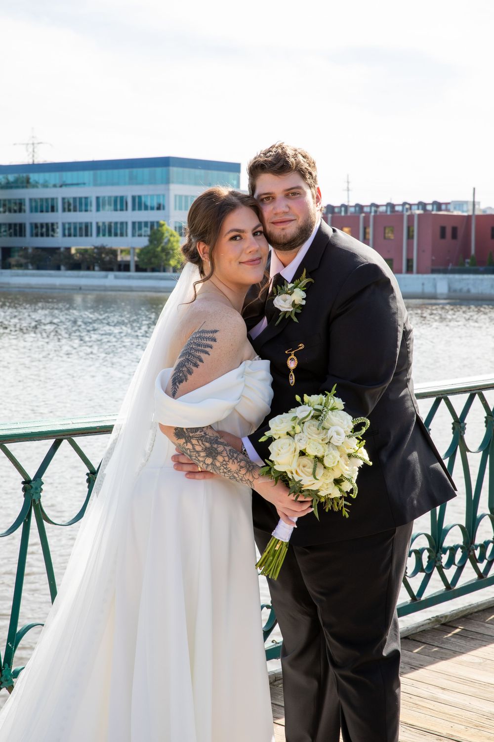 Newlyweds pose on a waterfront bridge, bride holding white bouquet in elegant wedding attire.