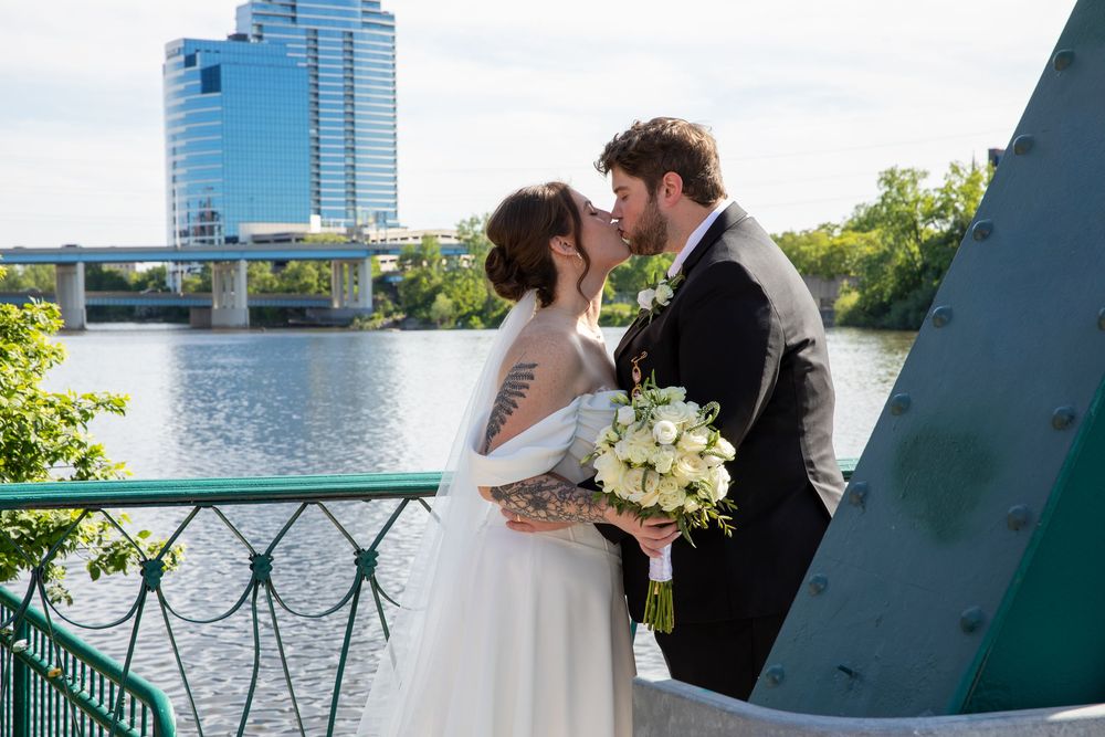 Bride and groom share a kiss on a bridge with river and city skyline in background.