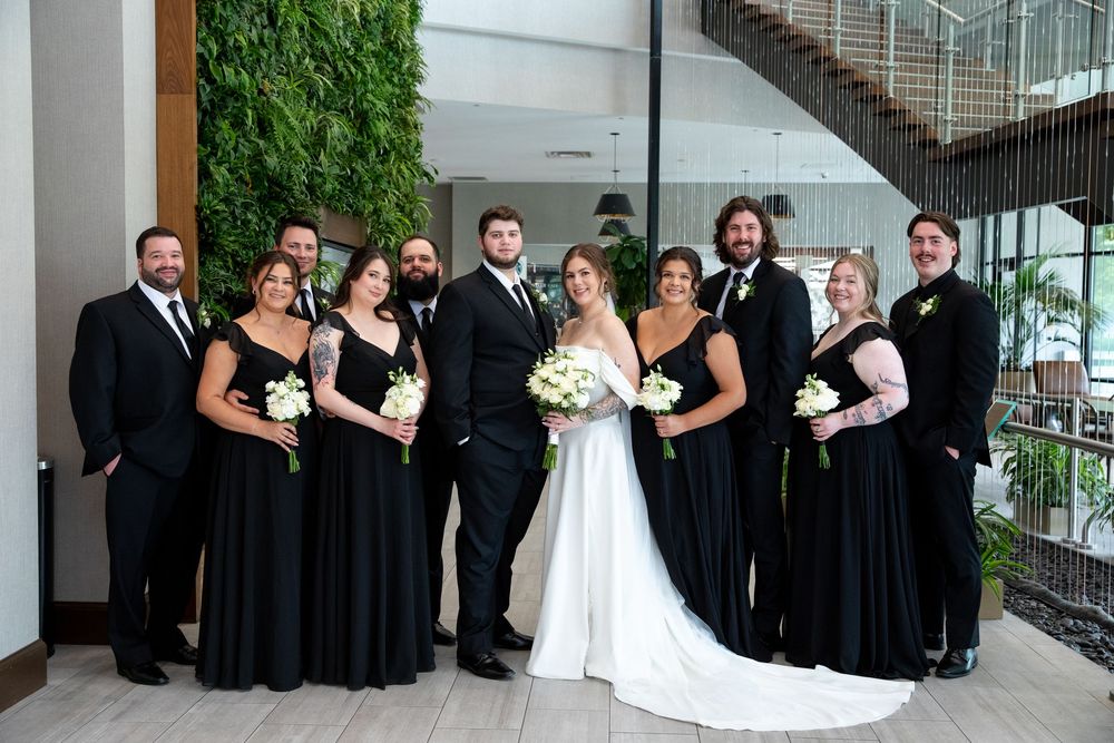 Wedding party of ten poses indoors, bridesmaids and groomsmen in black attire flanking bride and groom with bouquets.