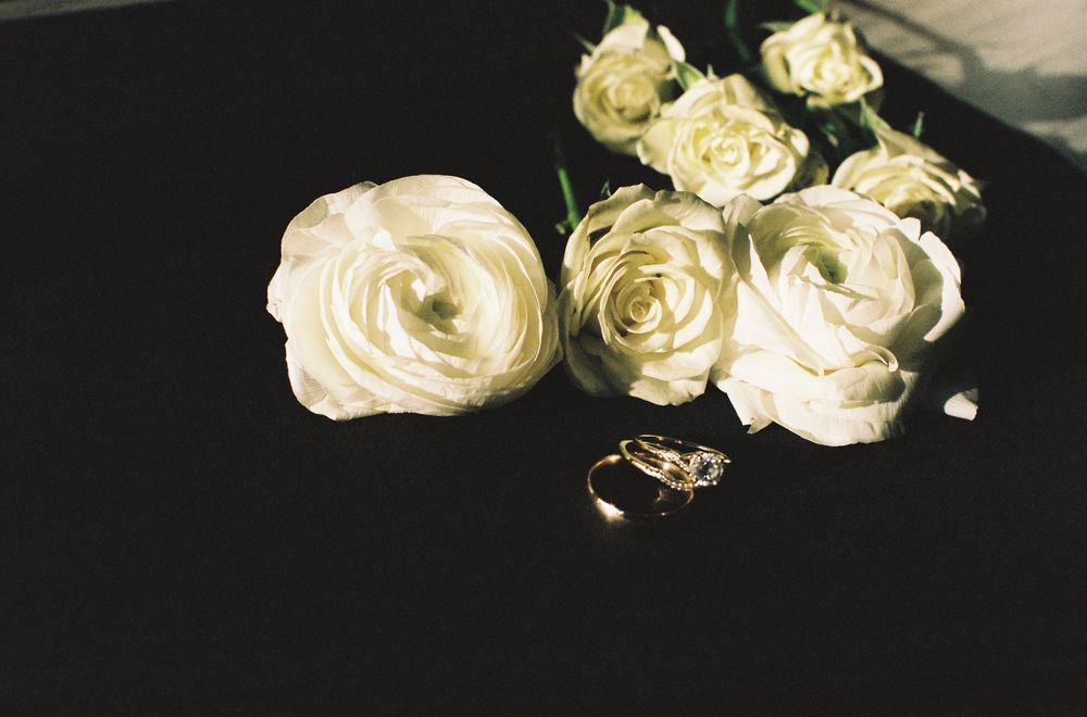White ranunculus and roses arranged on dark surface beside two gold wedding rings, elegant bridal flat lay.