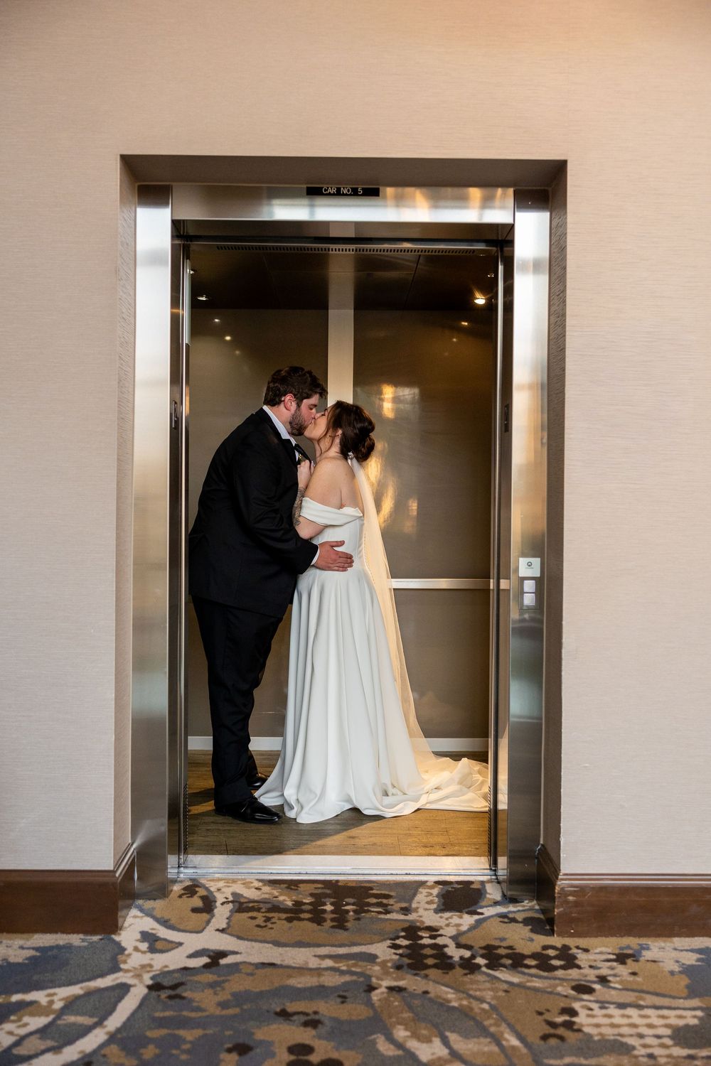 Bride and groom sharing a romantic kiss inside an elevator, bride wearing a flowing white gown with a long veil.