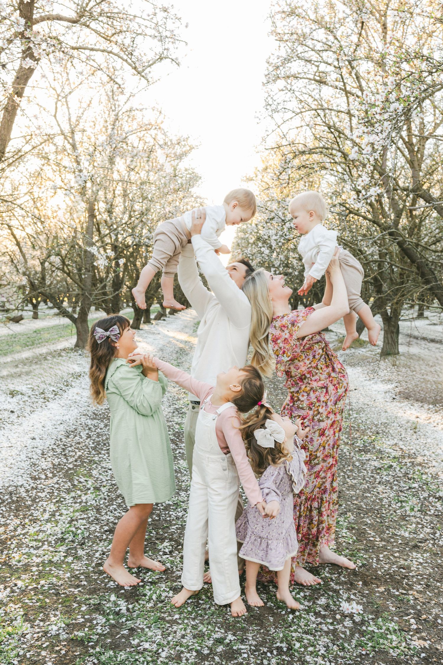 Family portrait in blooming orchard, parents lifting babies while children gather around, wearing pastel spring outfits.
