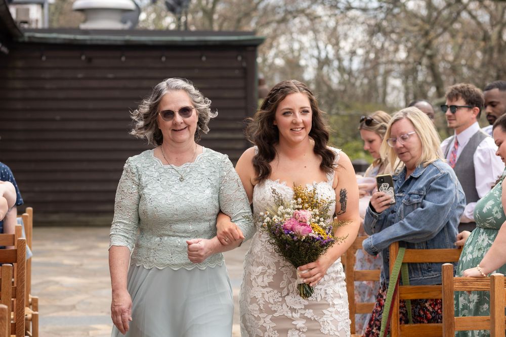 Bride walks down the aisle with her mother, holding a colorful bouquet at an outdoor wedding ceremony.