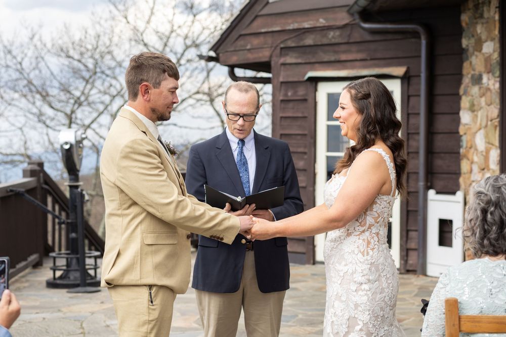 Couple exchanging vows outdoors at a rustic wedding ceremony with an officiant, bride in lace gown and groom in tan suit.