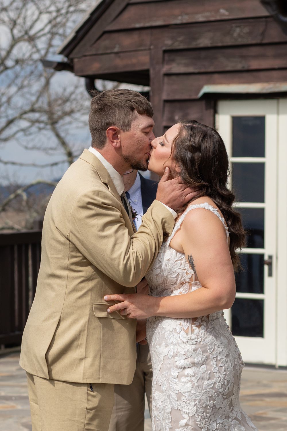 A bride and groom share their first kiss outdoors, wearing a white lace gown and tan suit with blue tie.