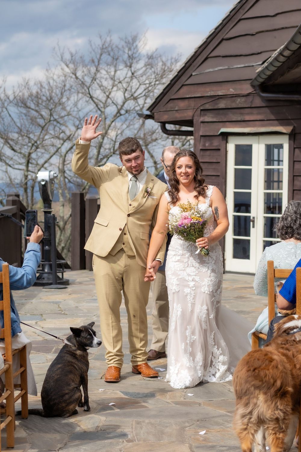 Newlyweds walk back down the aisle hand in hand, groom waving, with dogs watching at outdoor wedding.