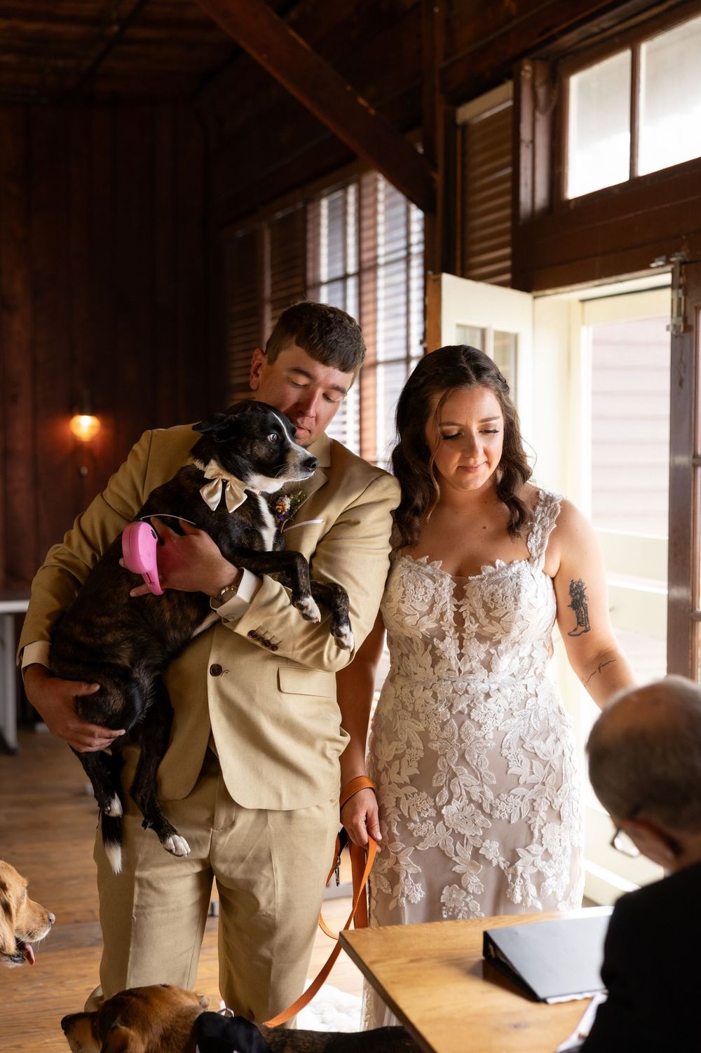 Bride and groom hold their dog dressed in a bow tie during their rustic indoor wedding reception.