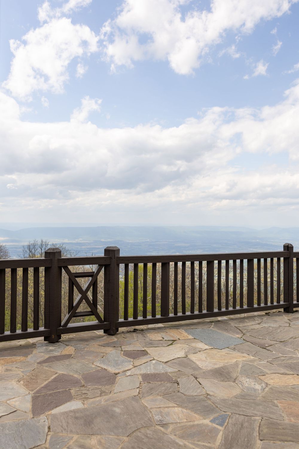 Stone patio overlook with wooden railing and panoramic mountain valley views under partly cloudy blue sky.