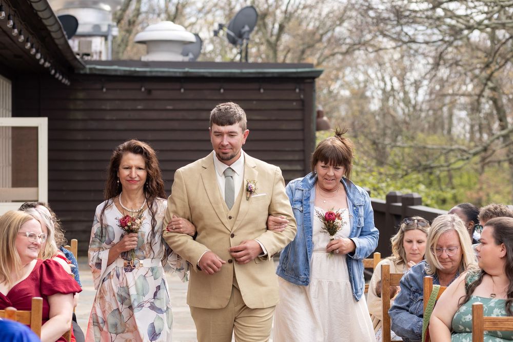 Groom in tan suit walks down outdoor wedding aisle escorted by two women holding pink flowers.