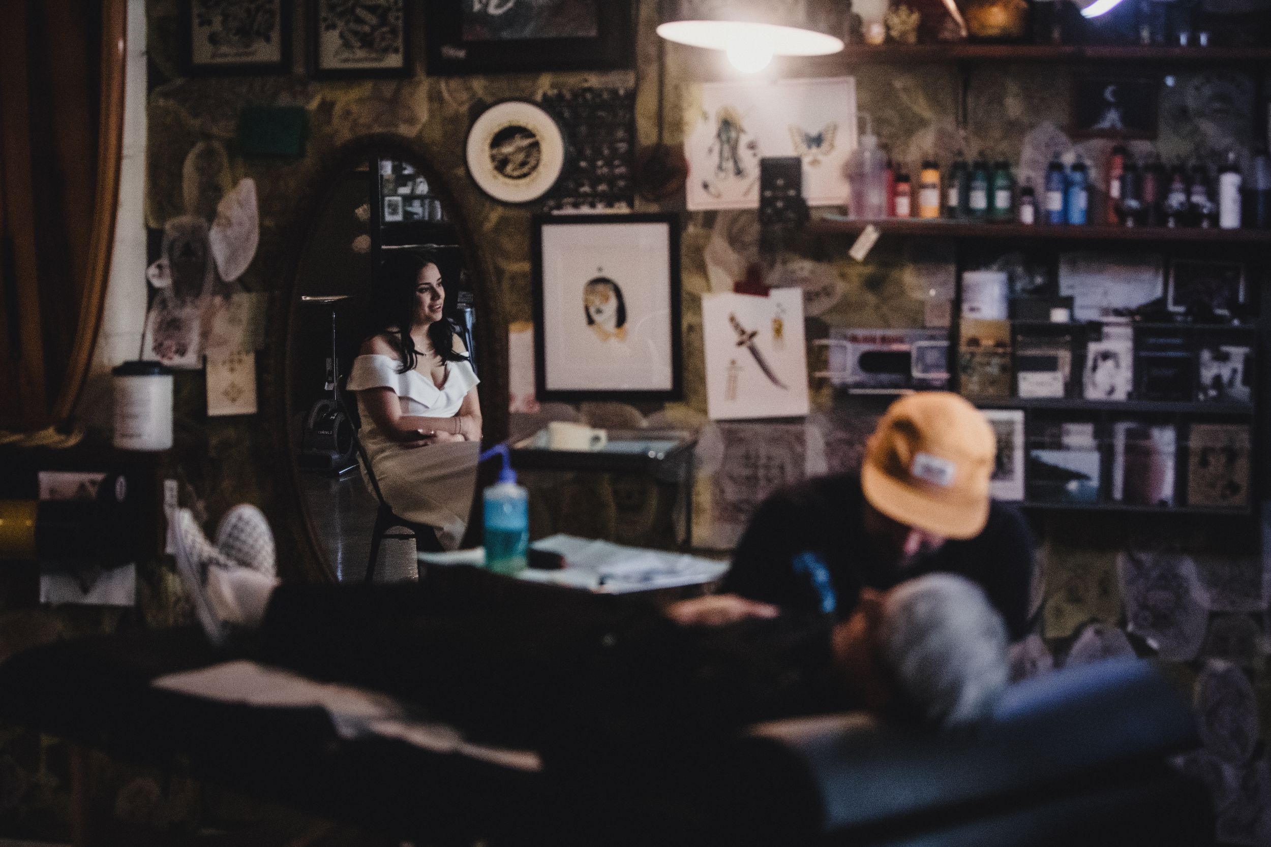 A tattoo artist works in a dimly lit studio filled with artwork and supplies while a client receives a tattoo.