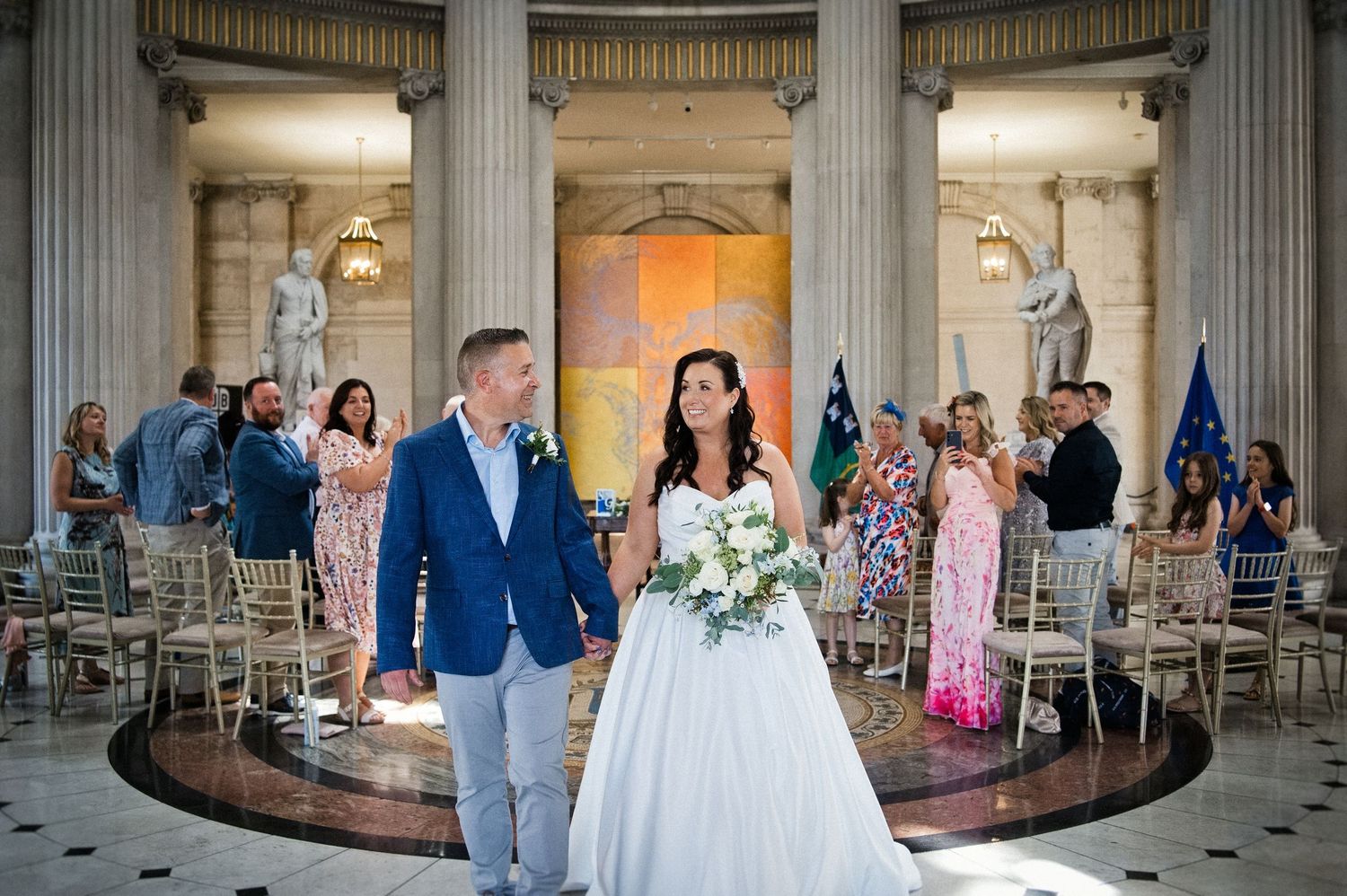 Bride and groom walk down the aisle in an elegant columned hall, guests seated on chiavari chairs during wedding ceremony.