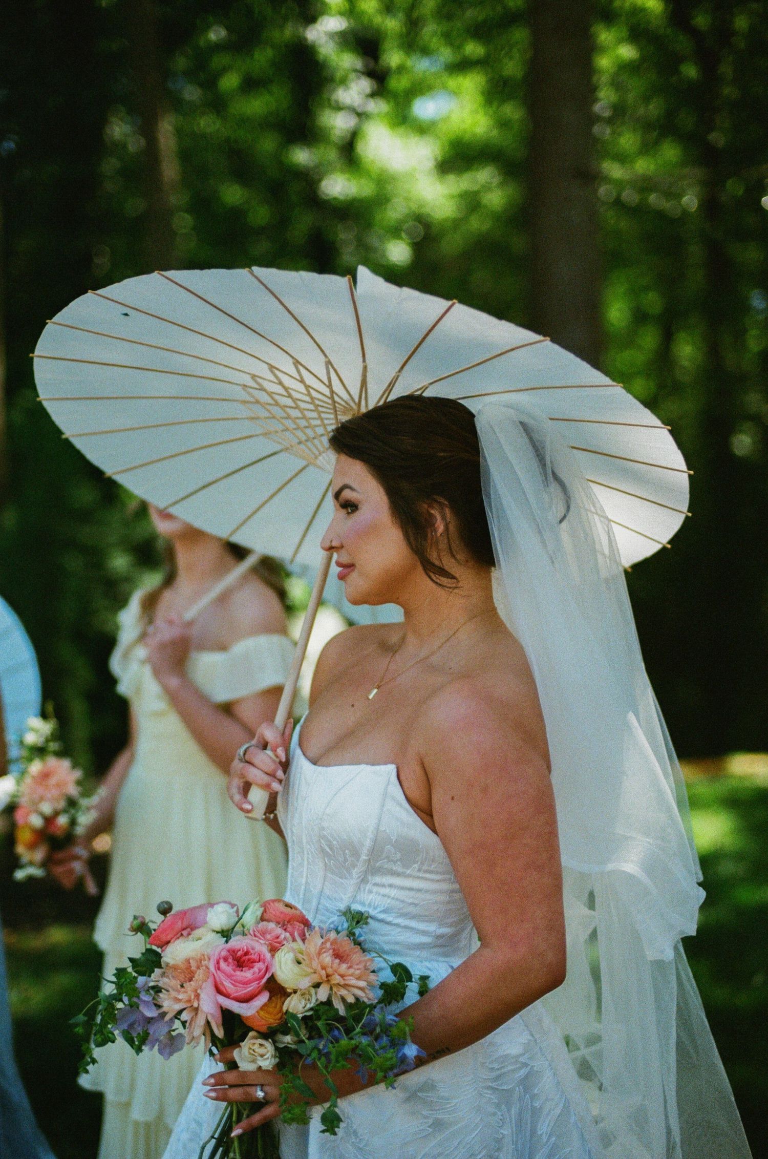 Bride in white gown holding parasol and colorful bouquet at outdoor wedding ceremony surrounded by trees.