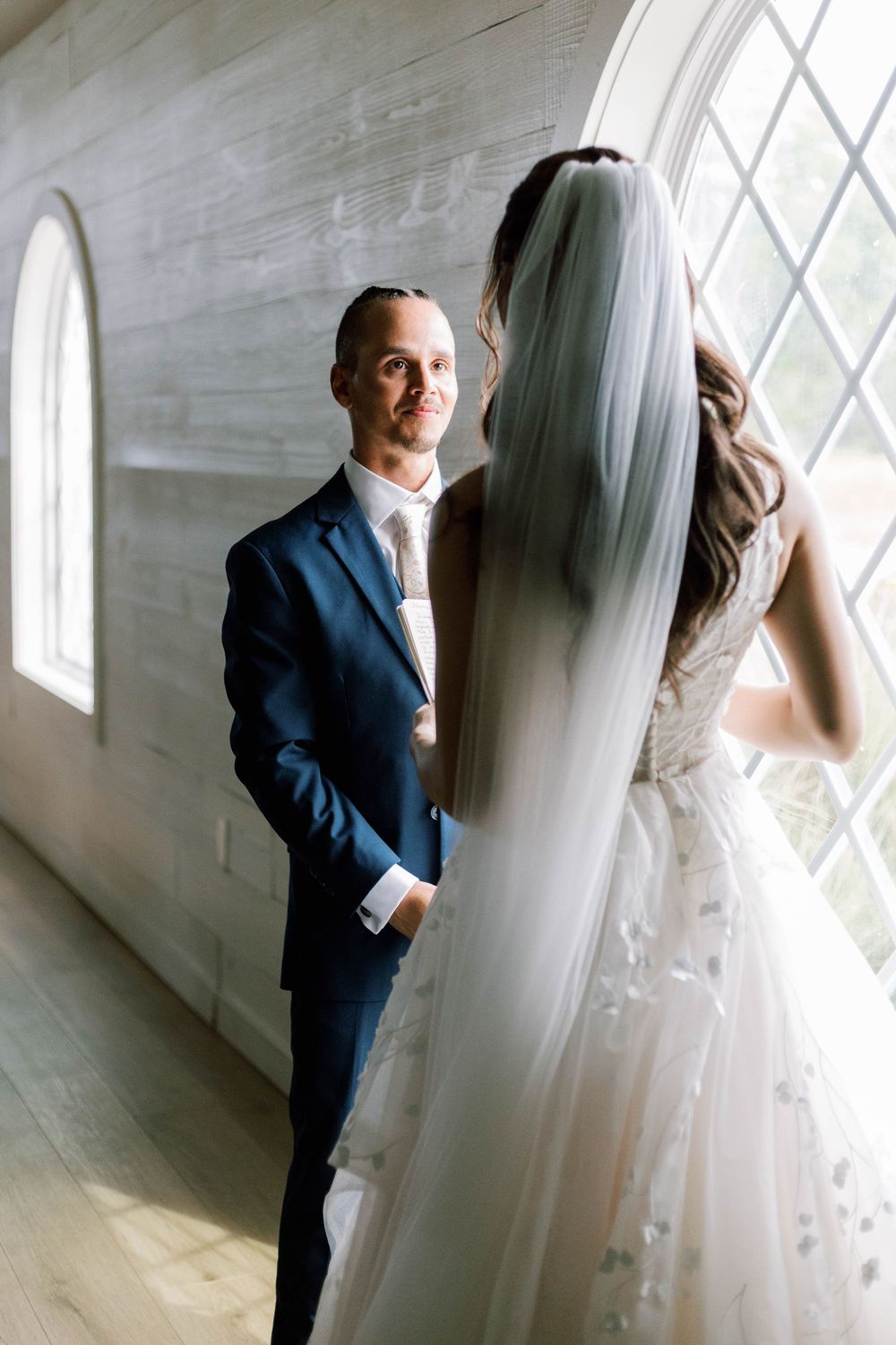 Groom in navy suit smiles at bride in white gown and veil standing by arched windows in elegant venue.