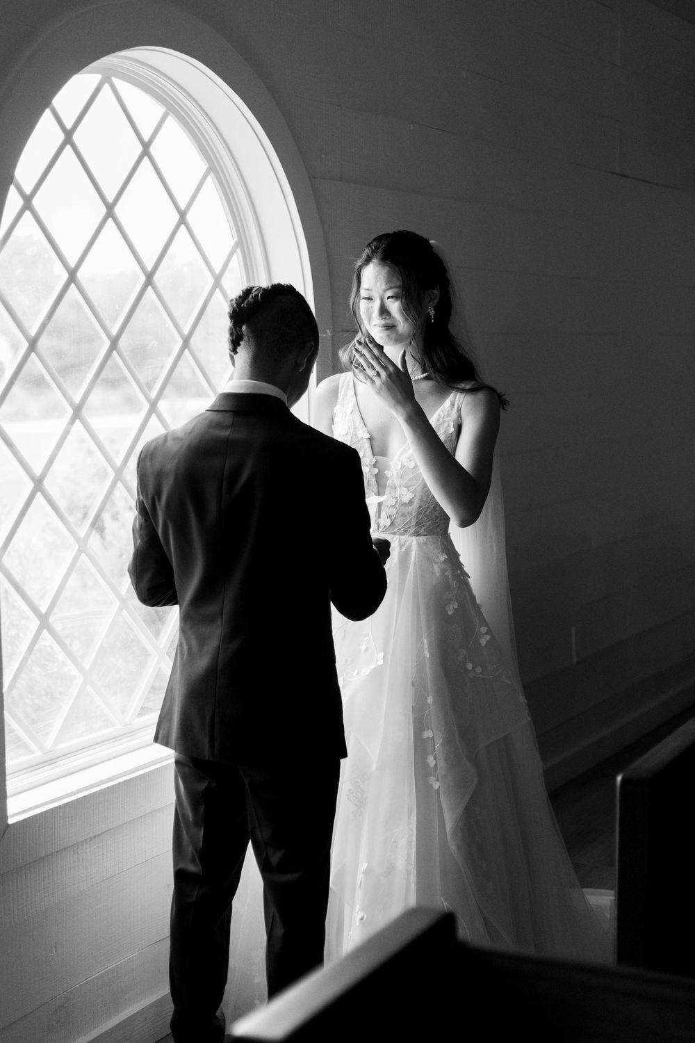 Black and white photo of a bride and groom sharing an emotional first look by an arched window in a chapel.