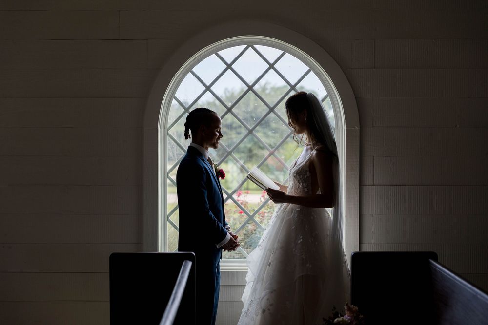 Bride and groom silhouetted against arched chapel window, exchanging vows in romantic backlit wedding ceremony photo.