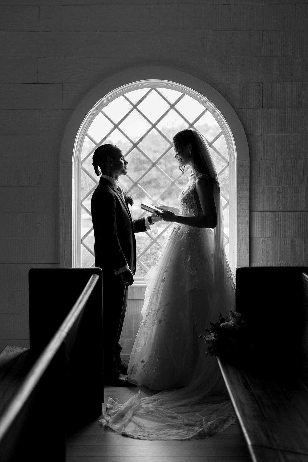 Black and white photo of bride and groom exchanging vows by arched church window, silhouetted in elegant wedding attire.