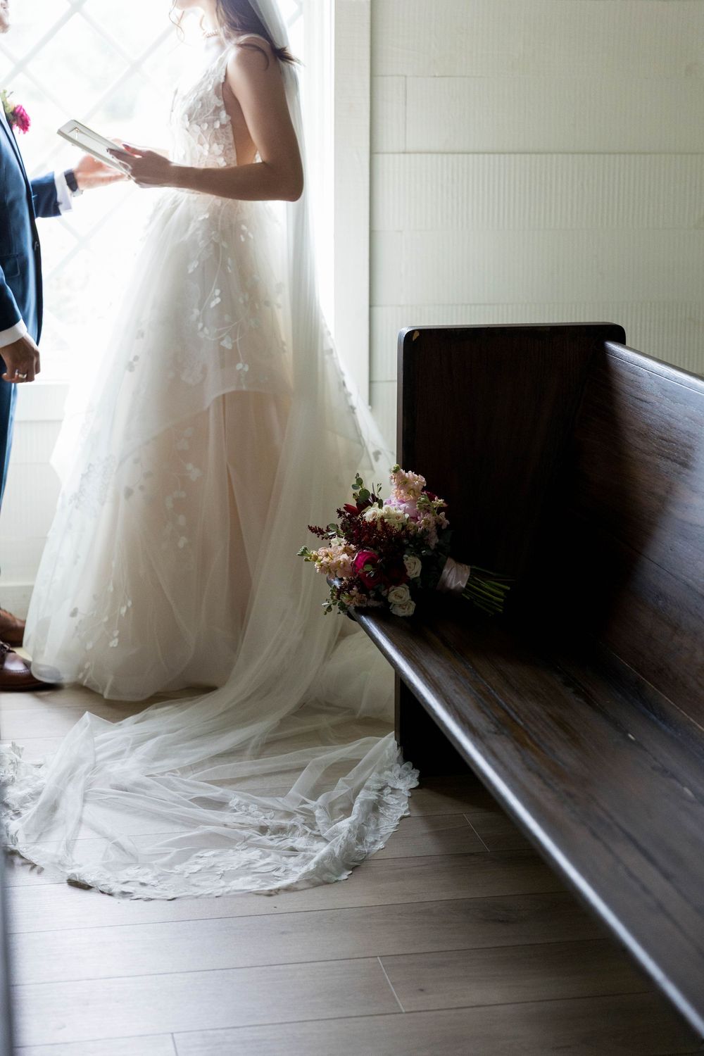 Bride in a white lace ball gown holding vows near a church pew with a floral bouquet resting beside her.