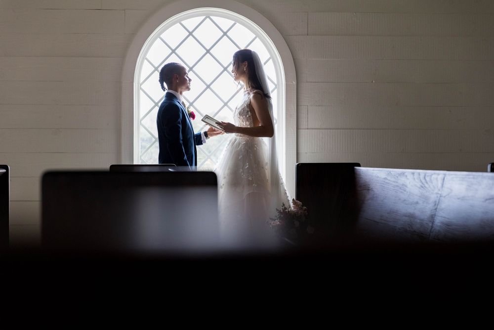 Bride and groom share an intimate moment by a arched window in a church, silhouetted by soft natural light.