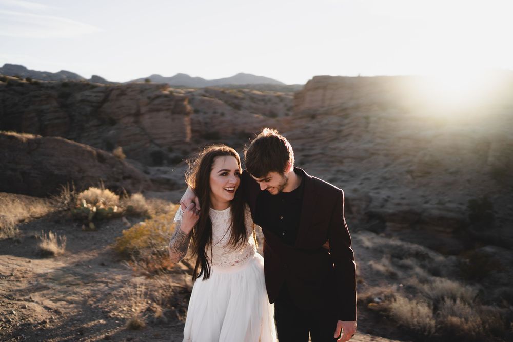 Bride in white dress and groom in dark suit walk together in golden sunset light among desert rock formations at San Lorenzo Canyon in NM