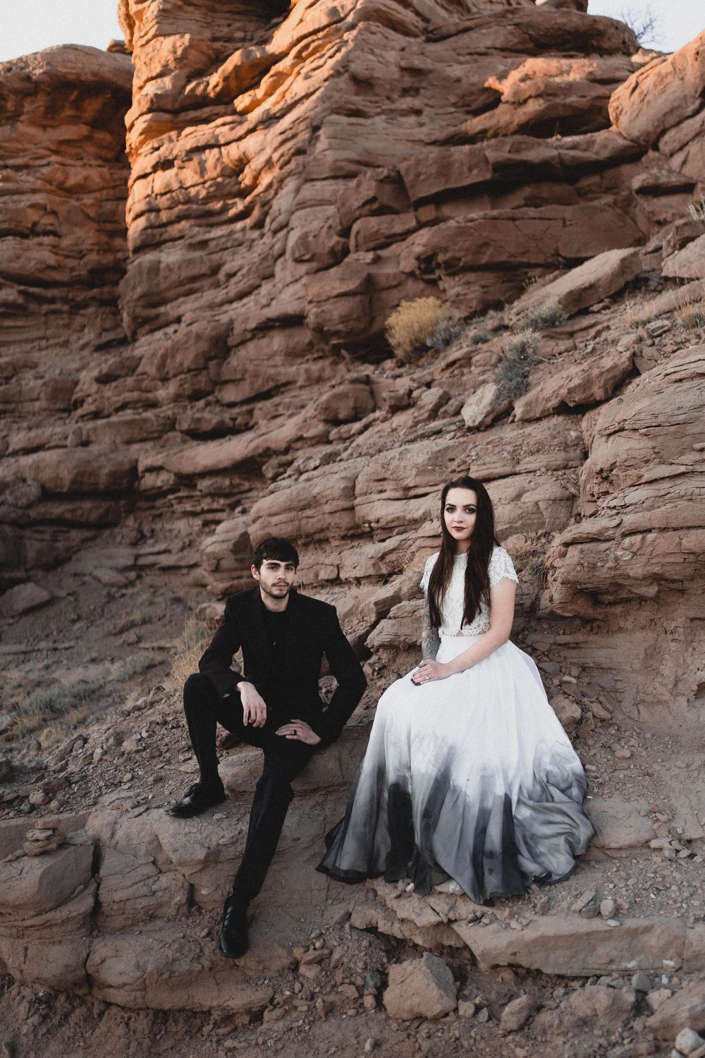 Bride in ombre white gown and groom in black suit pose seated on red sandstone rocks in San Lorenzo Canyon