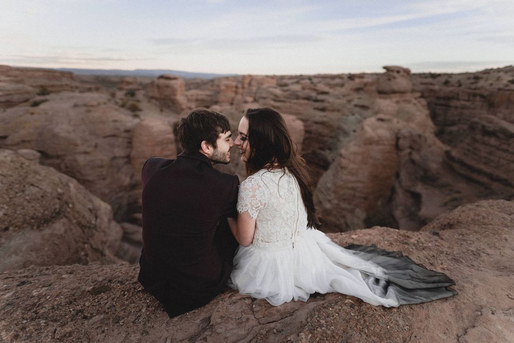 Bride and groom sit closely together sharing a kiss on red rock cliffs overlooking a vast desert San Lorenzo canyon.