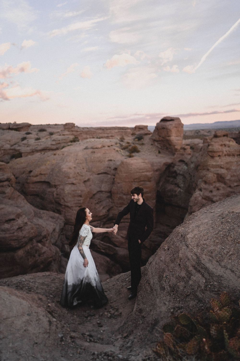 Bride in white and black ombre gown holds grooms hand among dramatic red rock formations at sunset at San Lorenzo Canyon