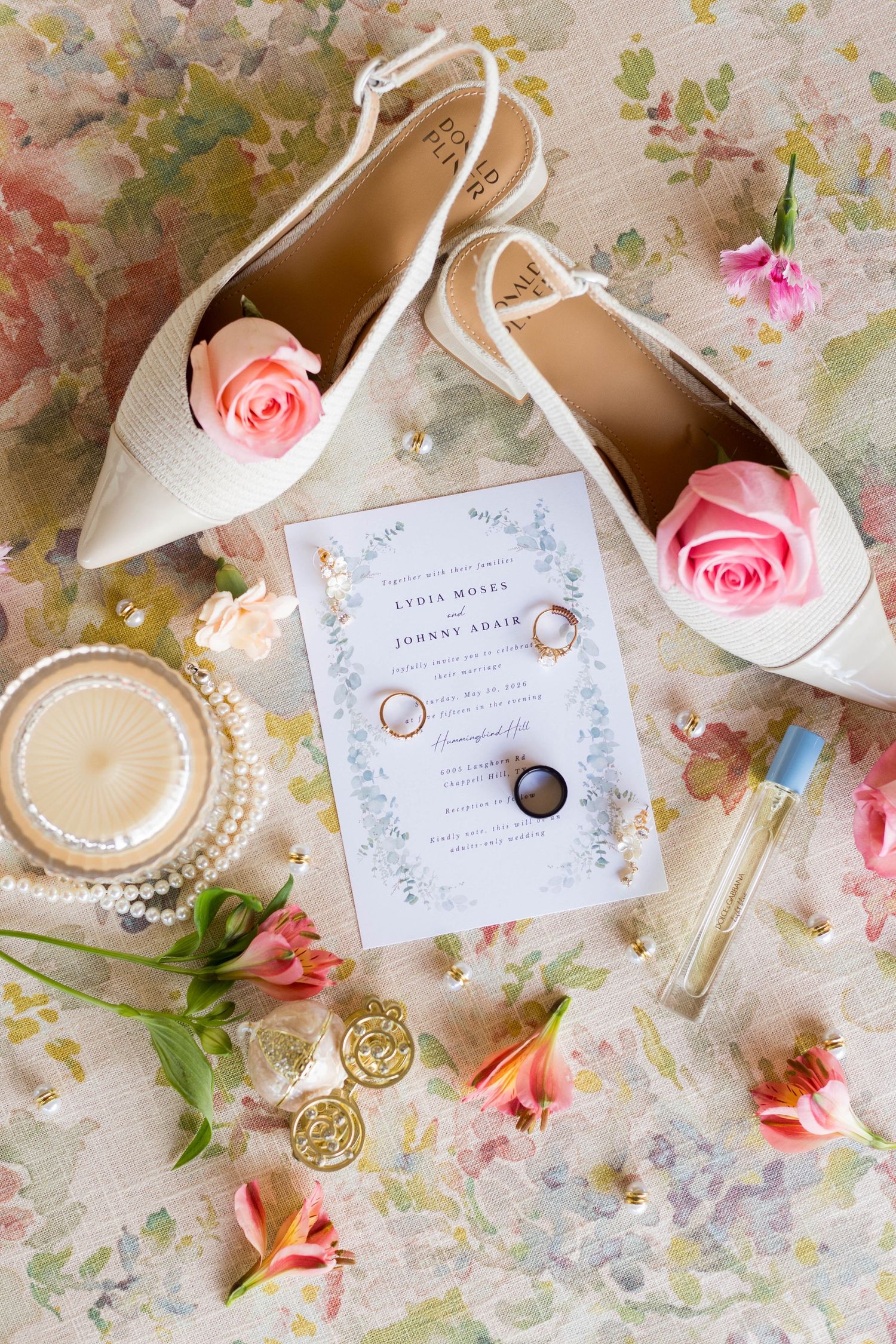 Elegant wedding flat lay with white heels, pink roses, gold rings, pearl bracelet, invitation, and floral accents on soft background.
