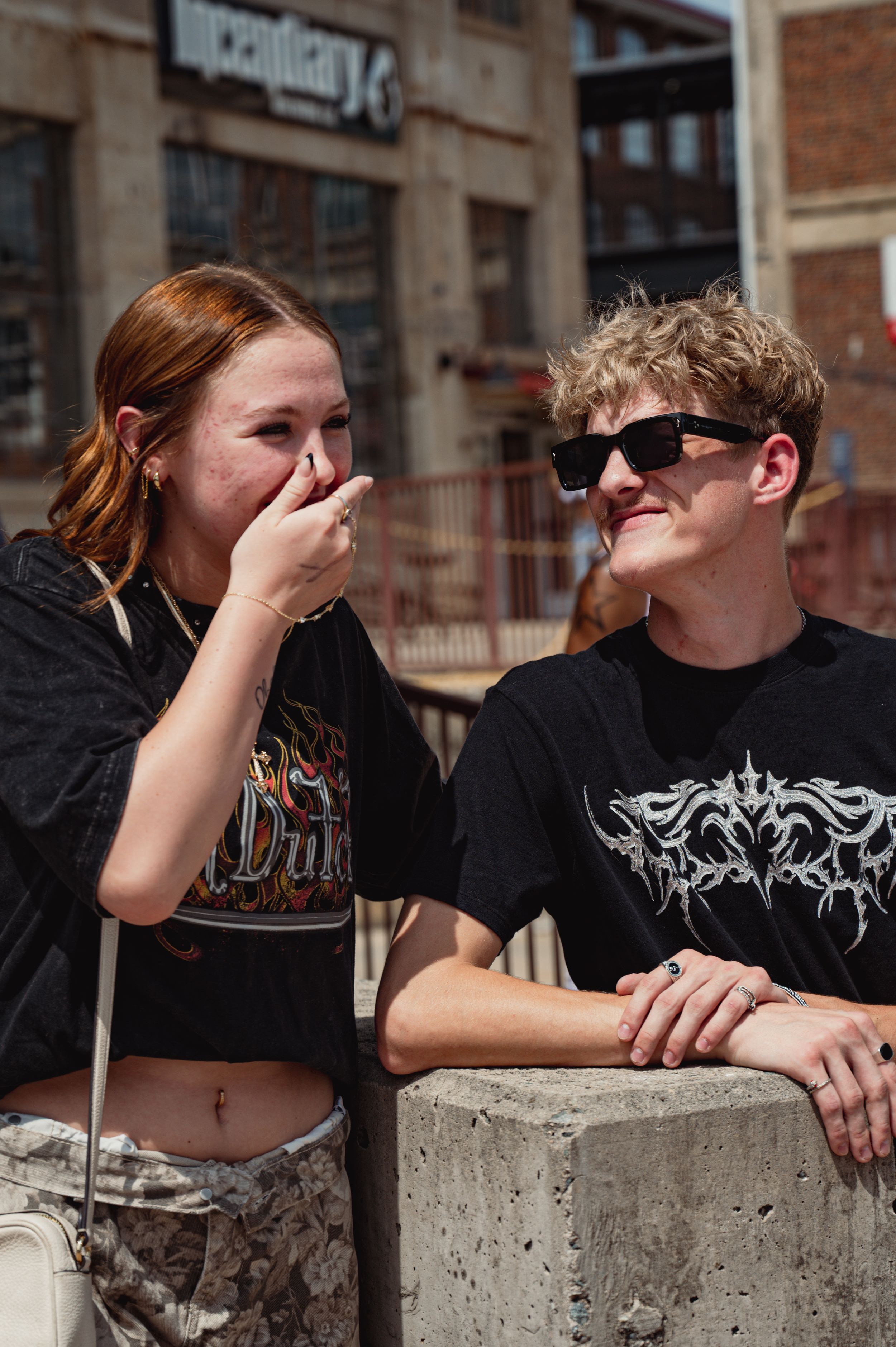 Two young friends laughing outdoors near urban building, girl in Duke tee covering mouth, boy in graphic tee wearing sunglasses.