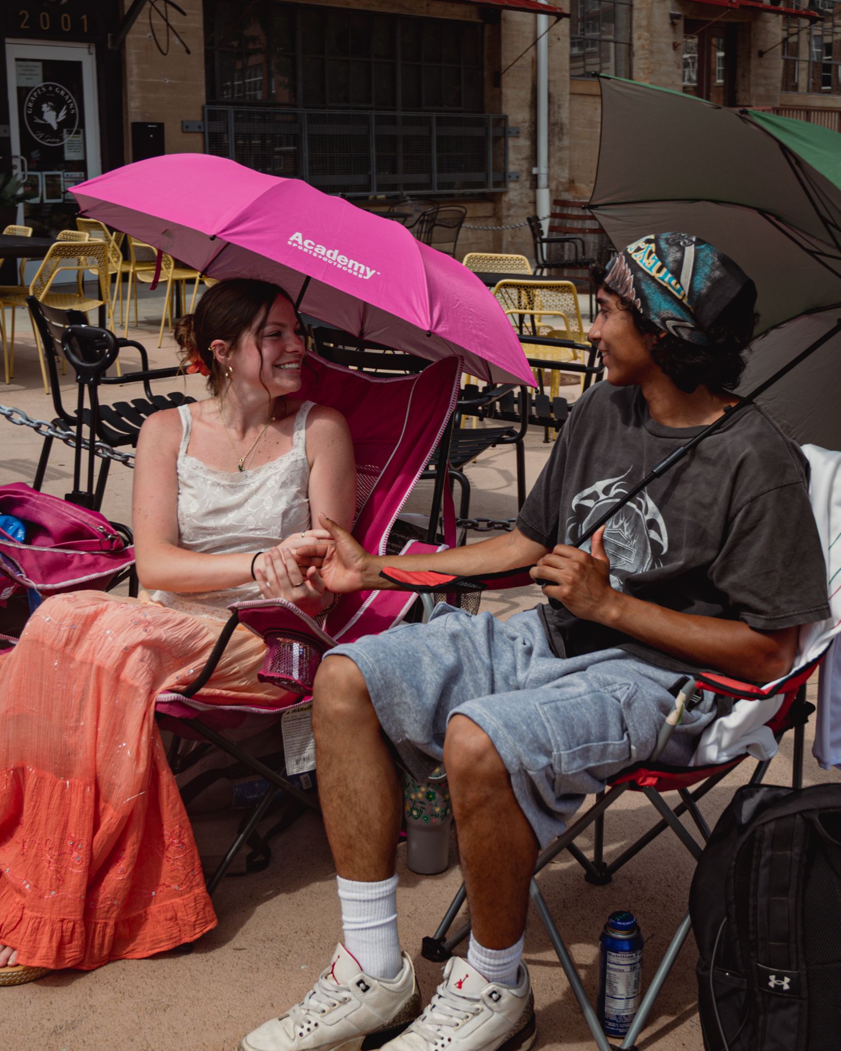 Two young people sitting in camping chairs under a pink Academy umbrella, holding hands and smiling outdoors.