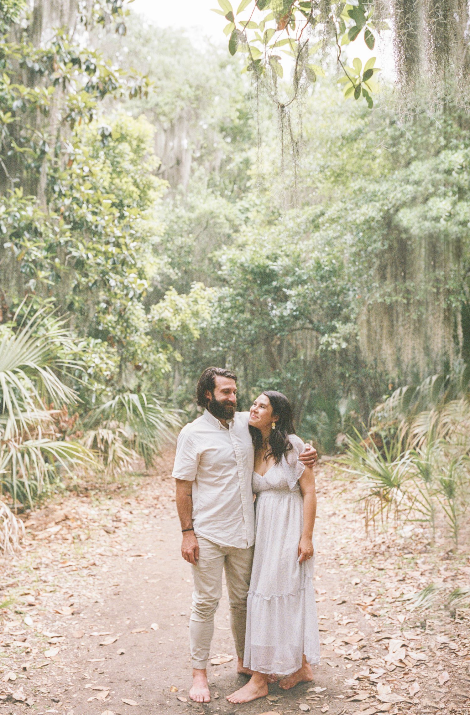 Couple standing on a forest path surrounded by lush greenery, woman in white dress leaning on bearded man in white shirt.
