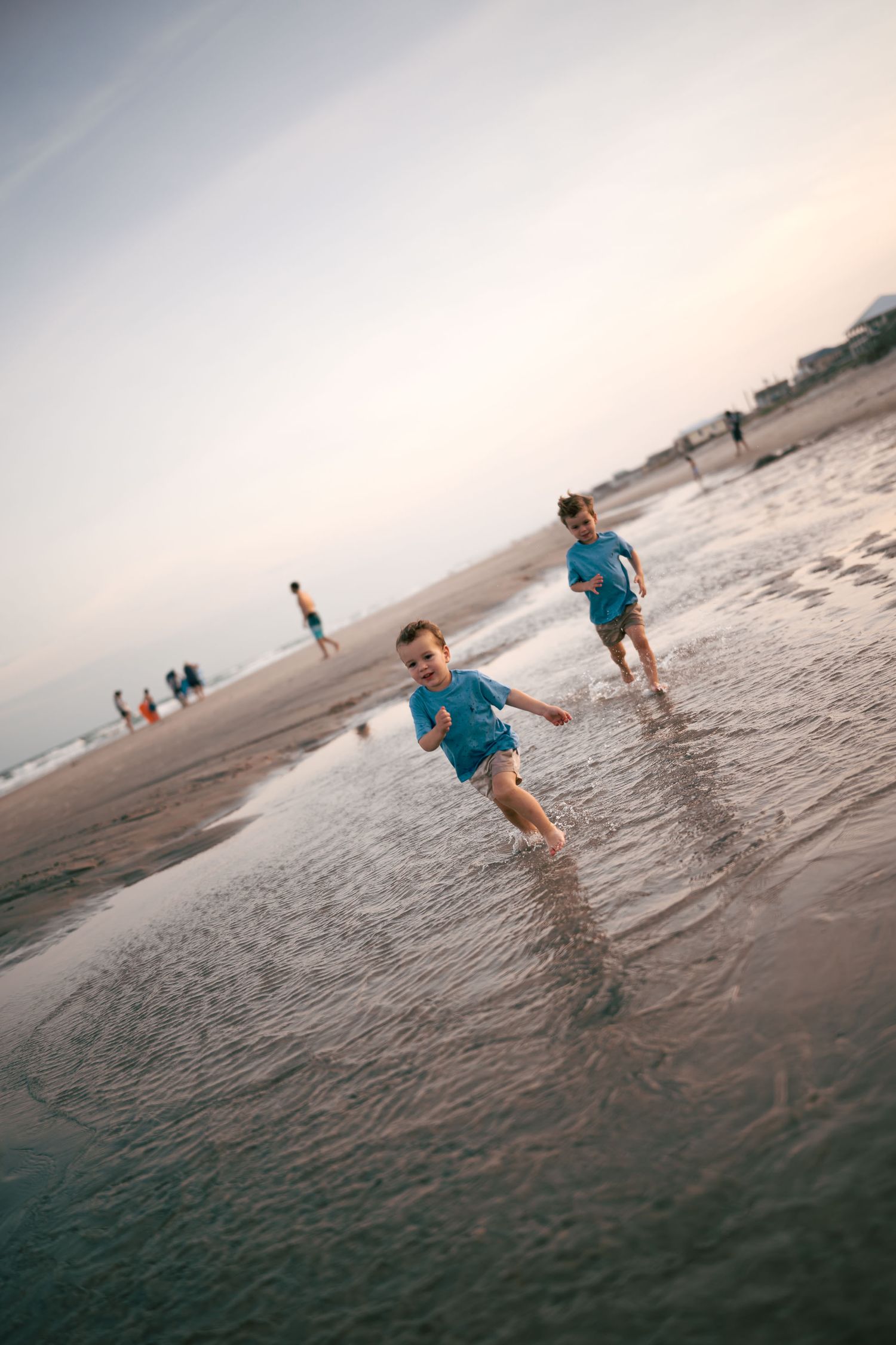 Two young children in blue shirts running through shallow ocean water at sunset.