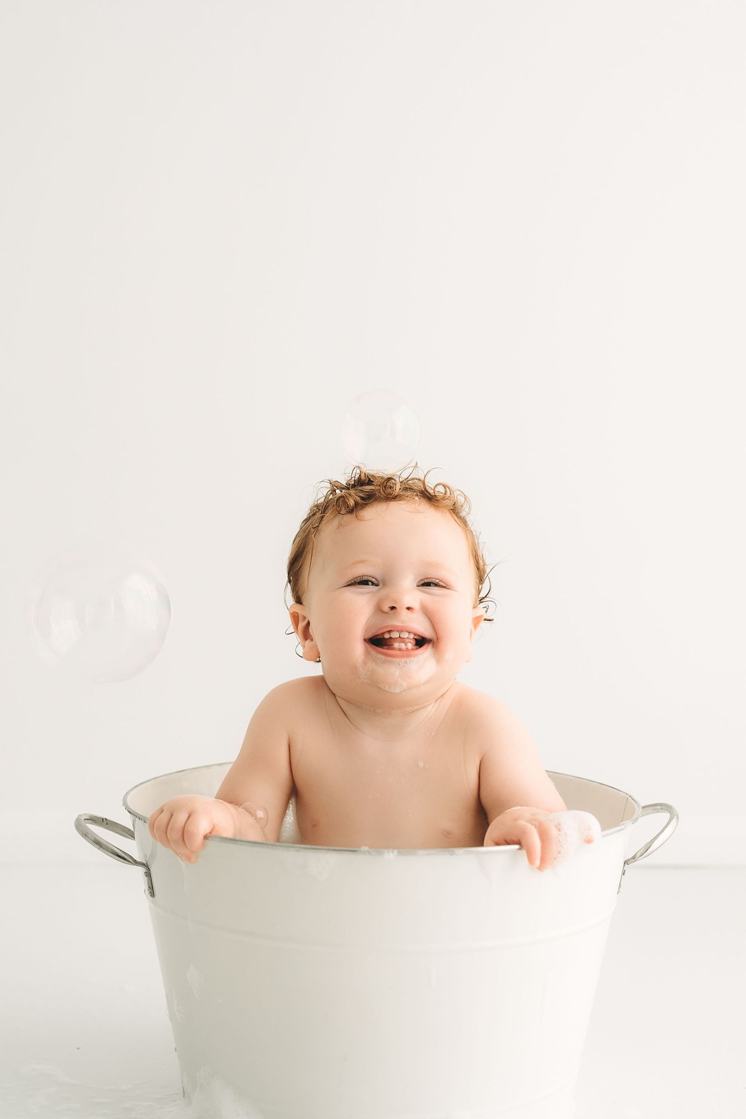 Happy laughing baby sitting in white metal tub with bubbles during bath time indoors.