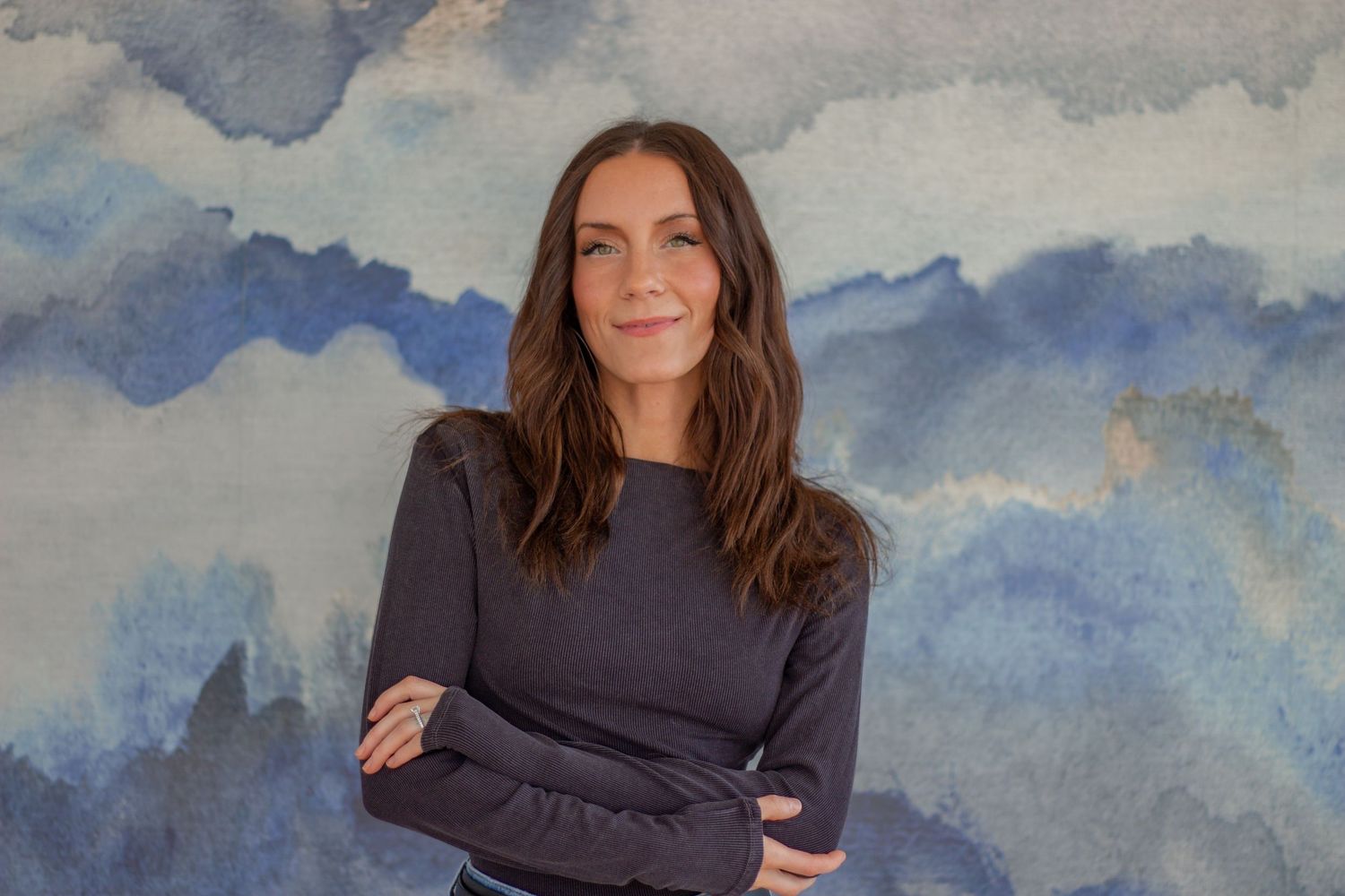 Woman with long brown hair smiling, arms crossed, wearing dark sweater against blue watercolor background.