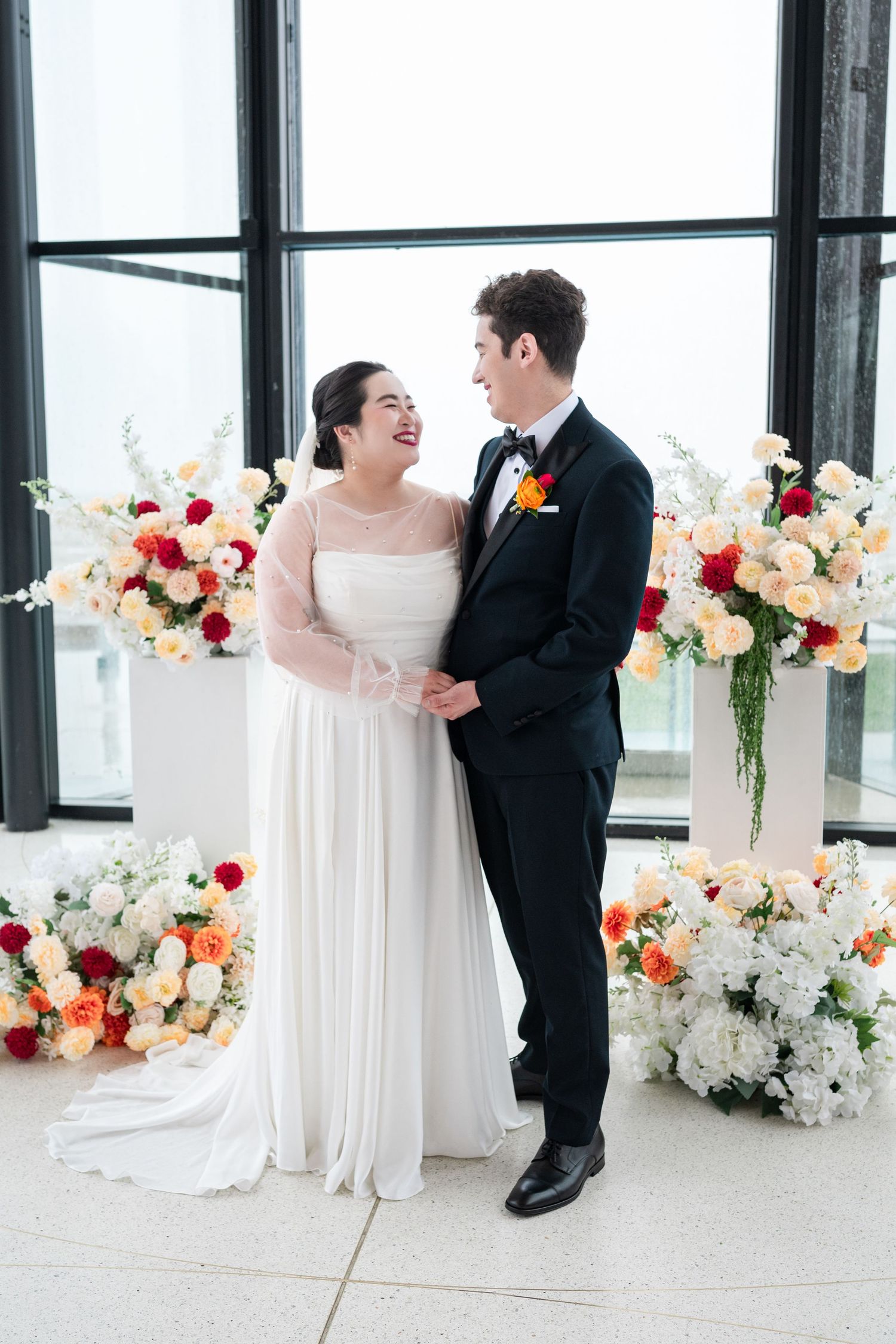 Joyful Asian bride in white gown with sheer sleeves holds notes and microphone, laughing during wedding speech.