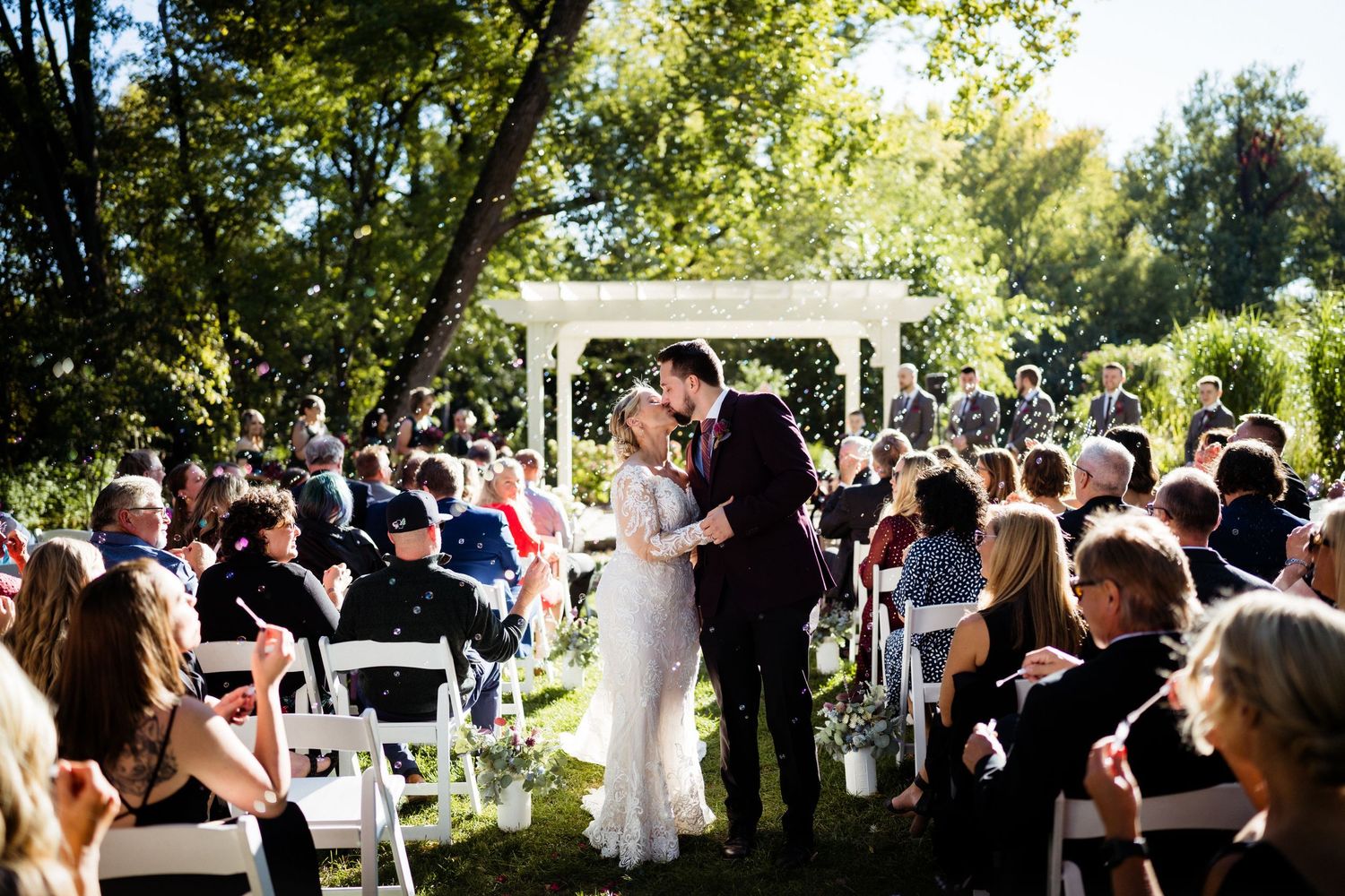 Bride and groom share first kiss at outdoor garden wedding ceremony with white gazebo and seated guests.