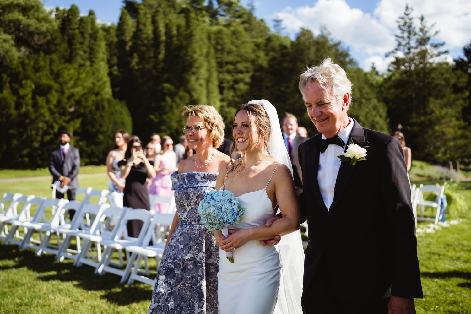 Bride and groom stand before guests in elegant white tent wedding reception with floral chandelier.