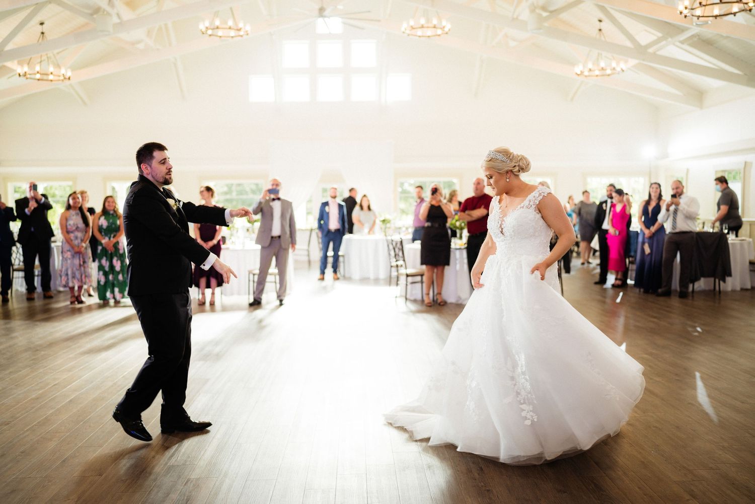 Bride and groom share their first dance in an elegant white reception hall surrounded by wedding guests.