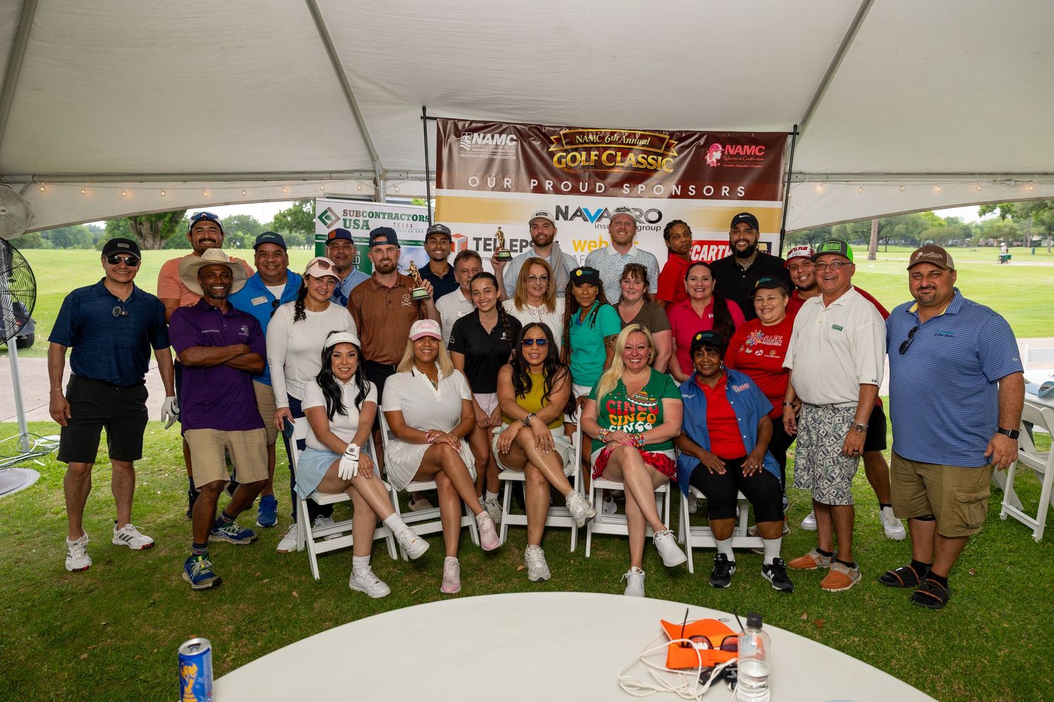 Large group of golfers posing under a tent at a charity golf classic tournament on a sunny day.