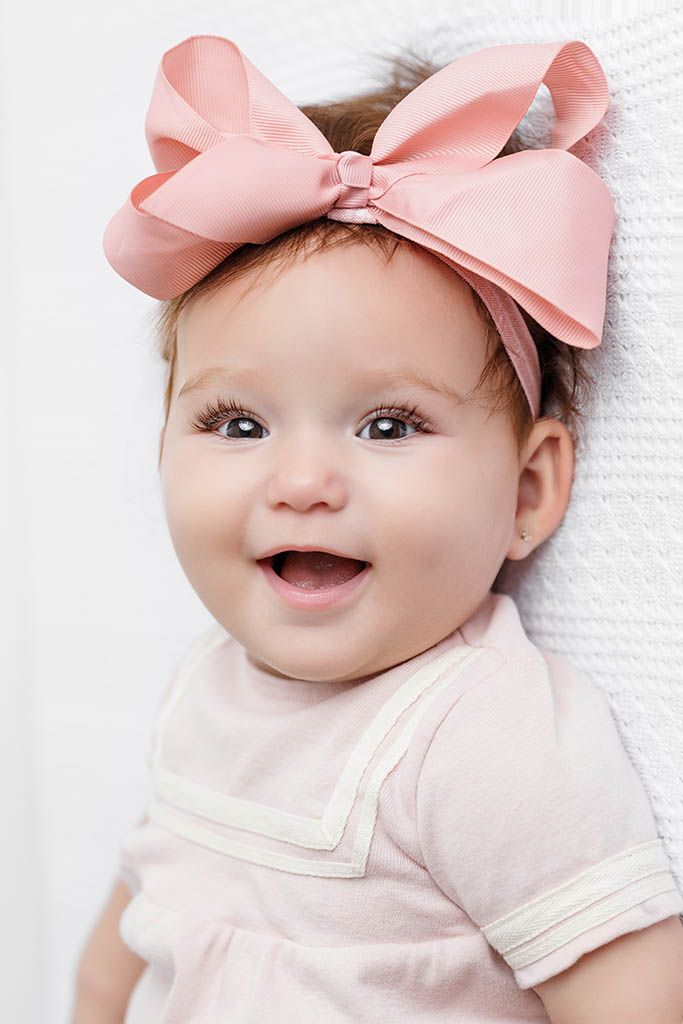 Smiling baby girl wearing a large pink bow headband and pink dress, with bright eyes and a joyful expression.