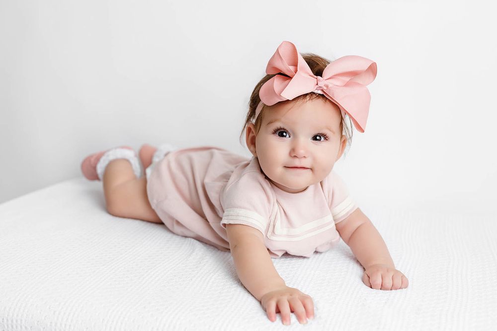 Adorable baby girl in pink romper and bow headband lying on white surface.