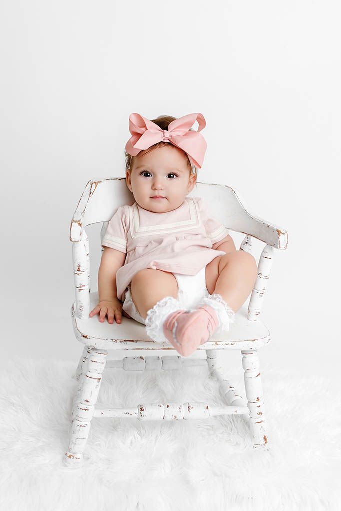 Baby girl in pink outfit and bow headband sitting in a white distressed rocking chair on a white background.