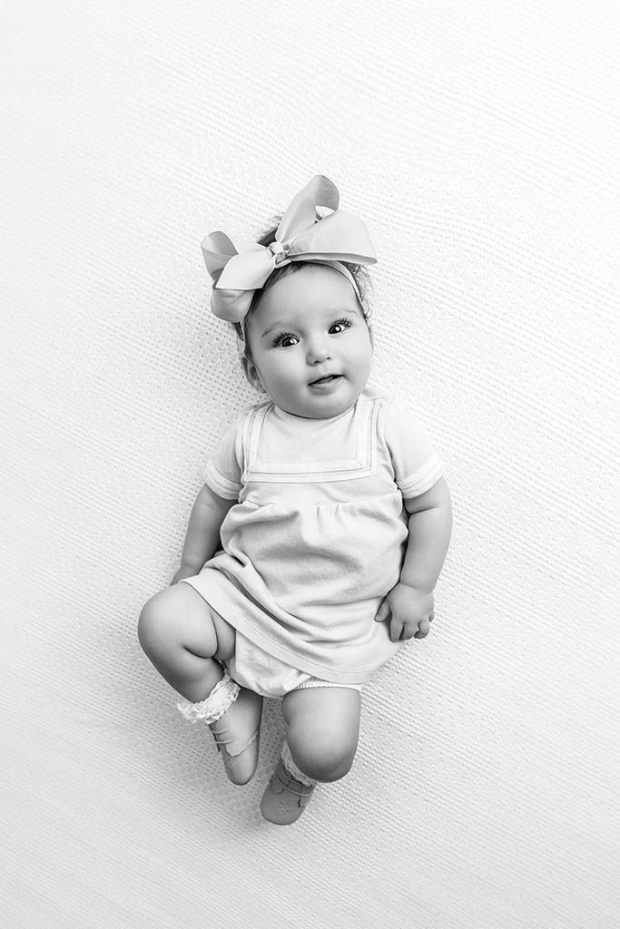 Black and white photo of a cute baby girl in a romper and bow headband lying on a white background.