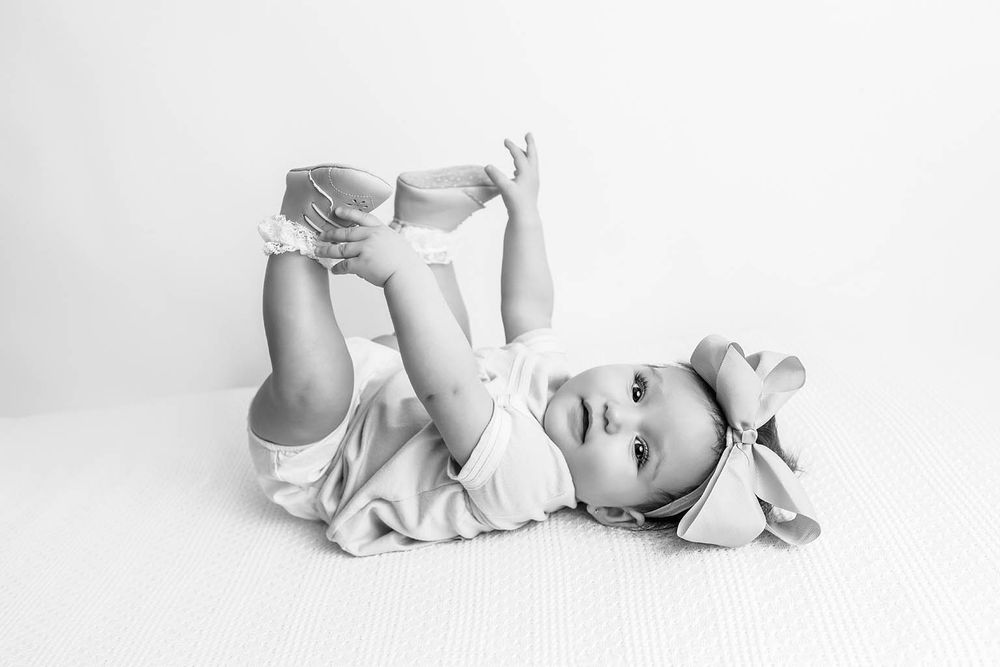Black and white photo of a cute baby lying on back, holding feet up, wearing a bow headband and dress.