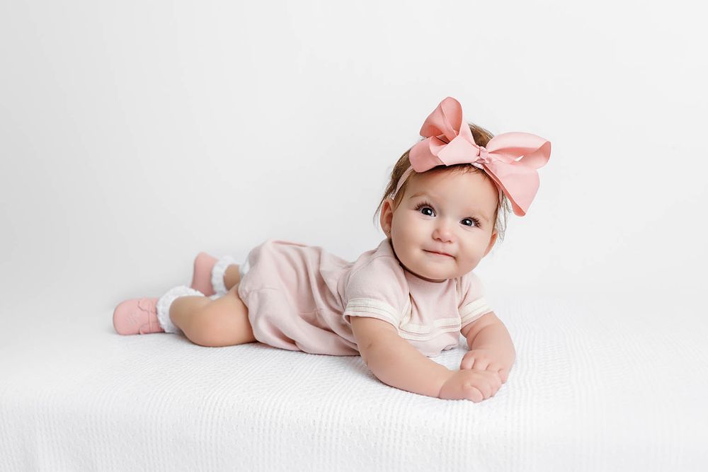 Cute baby girl wearing pink outfit and large bow headband lying on white bed.