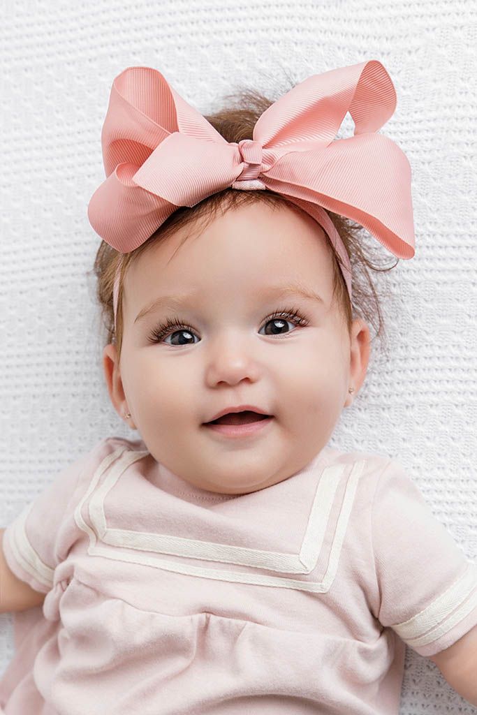 Adorable baby girl smiling, wearing a pink bow headband and pink dress, lying on a white textured surface.